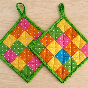 May include: Two square quilted pot holders featuring a patchwork design of small triangles in pink, orange, yellow, green, and blue, all patterned with white polka dots. Each pot holder is finished with a bright green border with a swirl pattern.
