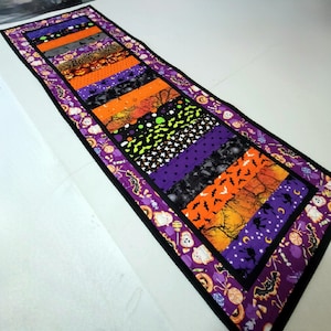 May include: A Halloween-themed table runner with a patchwork design. The runner features various fabrics with orange, purple, black, and gray patterns, including bats, pumpkins, and spooky trees. The edges are trimmed with a purple border.