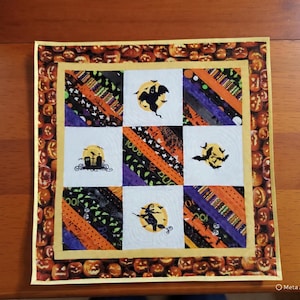 May include: A square fabric quilt featuring a border of orange pumpkins. The center grid contains four white squares with black silhouettes of a ghost, a graveyard, bats, and a witch on a broomstick, alternating with diagonal striped fabric panels.