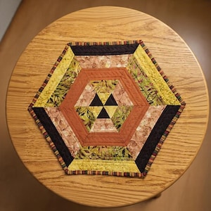 May include: A hexagonal quilted table mat featuring a central black triangle motif surrounded by concentric bands of fabric in shades of orange, yellow, and green with leaf patterns, finished with a dark brown border and a multi-colored striped edge.
