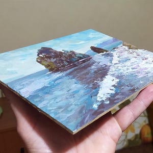 May include: A small painting on canvas depicting a rocky seascape with crashing waves. The painting is done in a realistic style with a blue and white color palette.