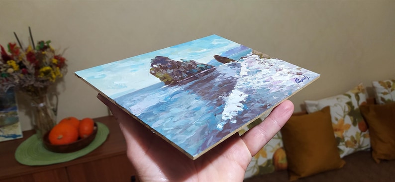 May include: A small painting on canvas depicting a rocky seascape with crashing waves. The painting is done in a realistic style with a blue and white color palette.