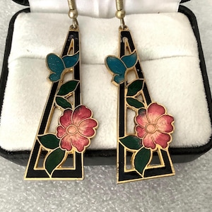 May include: A pair of dangle earrings featuring a black triangular frame with gold trim. Each earring has a pink flower, green leaves, and a teal butterfly. The earrings are displayed in a black jewelry box with a white interior.
