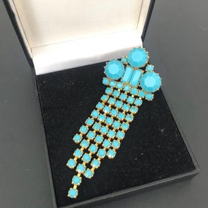 May include: A vintage turquoise and gold-tone brooch. The brooch features a cluster of large, round and rectangular turquoise stones at the top, with multiple strands of smaller turquoise stones cascading downwards. The brooch is set in a gold-tone metal and is displayed in a black box.