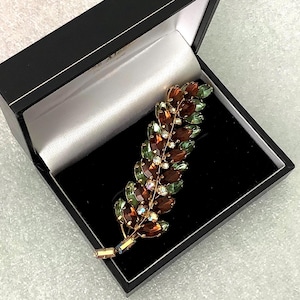 May include: A gold-coloured brooch shaped like a leaf, adorned with brown, green, and clear crystals. The brooch is presented in a black box with a white satin lining, offering a strong visual contrast.