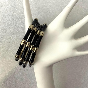 May include: A set of three black bangle bracelets featuring a segmented bamboo-style design with metallic gold-toned accents at each joint. The bracelets are stacked together, showcasing a glossy finish and a repeating pattern of black and gold.