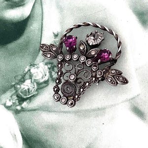 May include: A vintage silver-toned brooch in the shape of a basket, adorned with purple and clear gemstones. The basket handle is twisted, and the brooch features intricate filigree details and leaf accents, creating a classic, elegant design.