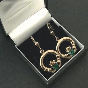 May include: Gold-tone Claddagh earrings with green gemstone accents, displayed in a black jewelry box with a white satin lining. The earrings feature a heart, crown, and hands design.