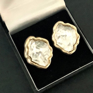 May include: A pair of white and gold-toned earrings in a black box. The earrings have an irregular shape with a gold-colored border. The center of each earring is white with a textured, crystalline appearance. The earrings are displayed in a black jewelry box.