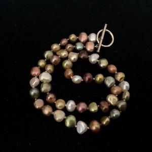 Könnte beinhalten: Eine mehrsträngige Halskette mit unregelmäßigen Barockperlen in verschiedenen schillernden Farben wie Bronze, Olivgrün, Creme und zartem Rosa. Die Kette wird mit einem polierten Knebelverschluss aus goldfarbenem Metall geschlossen.