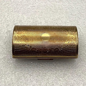 May include: A rectangular gold-toned metal case featuring an intricate engine-turned crosshatch pattern across the surface. The lid is decorated with a delicate floral border and a smooth, circular central medallion for engraving or personalization.