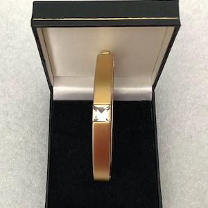 May include: A gold-toned hinged bangle bracelet with a matte finish, featuring a single square-cut clear gemstone set in the center. The bracelet is displayed upright inside an open black jewelry box with a white interior lining and gold trim.
