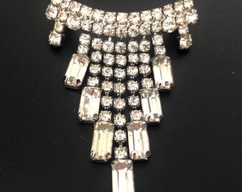 May include: A silver-toned brooch featuring a curved top bar encrusted with small round clear rhinestones. Below the bar, five vertical strands of round rhinestones descend, interspersed with larger rectangular clear baguette-cut rhinestones.