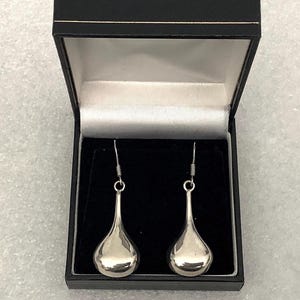 May include: A pair of polished silver-toned drop earrings with a smooth, teardrop-shaped design. The earrings feature thin metal hooks and are presented inside a black rectangular jewelry box with a white satin-lined interior lid.