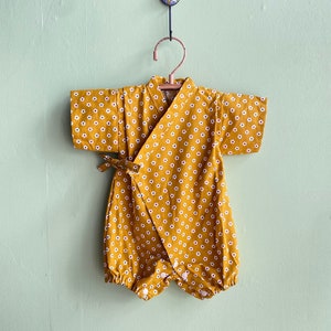Könnte beinhalten: Ein gelber Baby-Strampler mit weißem Blumenmuster. Der Strampler hat einen Wickelverschluss und hängt an einem Holzbügel.