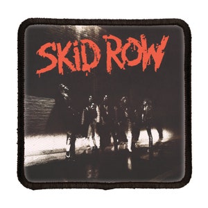 May include: A black and white band patch with the text "SKID ROW" in red lettering. The band is standing in a dark alleyway.