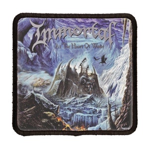 May include: A black and white patch with the band name "Immortal" and the album title "At The Heart Of Winter" in a silver font. The image features a dark, icy landscape with a fortress in the center.