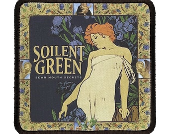 May include: A square patch with a black border and a vintage-style design. The patch features a woman in a white dress with red hair, surrounded by blue irises. The text "Soilent Green" is printed in gold letters with the words "Sewn Mouth Secrets" below.