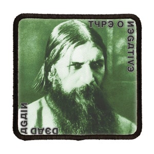 May include: A green and white patch with a black border. The patch features a black and white photo of a man with a beard and long hair. The text "dead" and "AGDII" are printed in black on the left side of the patch. The text "NEGATIV" and "3 O ЧУР" are printed in black on the right side of the patch.