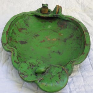 May include: A green, frog-themed decorative dish. The dish is shaped like a lily pad with a frog figurine perched on the edge. The dish has a distressed finish, with brown accents. The dish is designed to hold small items.