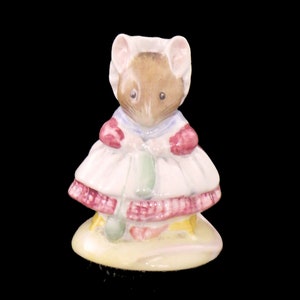 May include: A ceramic figurine of a mouse wearing a white dress with a pink and red trim, a blue collar, and a white bonnet. The mouse is standing on a yellow base against a black background.