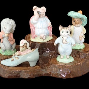 May include: A collection of ceramic figurines featuring animal characters. The figurines include a squirrel, a pig with a monocle, a cat, a rabbit, and a mouse in a shoe. The figurines are set on a brown, tree-stump-shaped base.