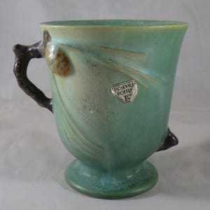 Può includere: Una tazza vintage in ceramica verde con un manico a forma di ramo e un piccolo dettaglio a forma di pigna. La tazza ha una forma svasata e una base a piedistallo. La tazza ha un adesivo con la scritta "Roseville Pottery".