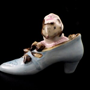 May include: A ceramic figurine of a mouse wearing a pink and white checkered shirt and a white hat with a pink stripe, peeking out of a light blue high-heeled shoe. The shoe has brown accents and a decorative design on the toe.