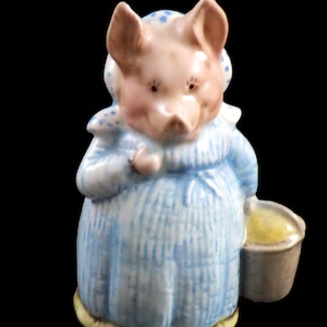 May include: A ceramic figurine of a pig wearing a blue dress with vertical stripes and a matching bonnet with blue polka dots. The pig is holding a small bucket with a yellow substance. The figurine has a light pink face and ears.