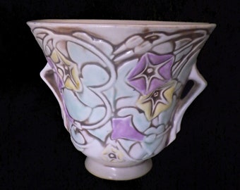 May include: A white ceramic planter with a floral design. The planter has two handles and a scalloped rim. The flowers are in shades of purple, yellow, and green.
