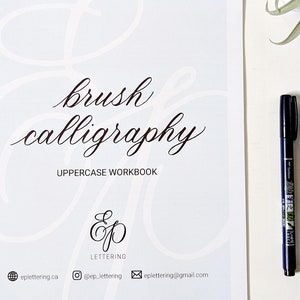 Op de afbeelding: Een wit werkboek met de titel "brush calligraphy uppercase workbook" en een logo voor "ep lettering". Twee zwarte en blauwe pennen liggen op een wit oppervlak naast het werkboek.