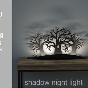 Puede incluir: Una silueta de metal negro de tres árboles con un búho posado en uno de los árboles. La silueta está iluminada desde atrás, proyectando una sombra sobre una superficie de madera. El texto "shadow night light" está impreso debajo de la silueta.