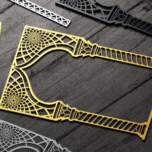 May include: A gold decorative frame with intricate geometric patterns and a rope-like design. The frame is made of thin metal and has a cutout design. The frame is on a dark wood background.