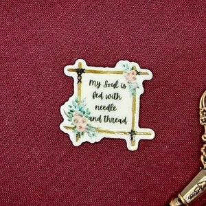 Op de afbeelding: Een bordeauxrode stof gespannen over een houten borduurring met een bloemensticker waarop staat "Mijn ziel wordt gevoed met naald en draad". Een gouden schaar met sierlijke handvatten ligt op de ring.