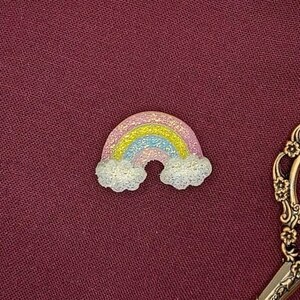 May include: An embroidery hoop with a glittery rainbow and clouds design on a maroon fabric background. The hoop is surrounded by a spool of thread and a pair of ornate scissors.