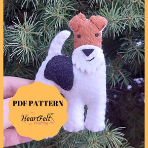 Puede incluir: Patrón PDF para un adorno de perro fox terrier de pelo duro de fieltro. El perro es blanco con marcas negras y marrones. El patrón está disponible para descargar en HeartFelt Crafting Co.