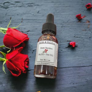 May include: A clear glass bottle with a black dropper top filled with a rose-colored liquid. The bottle has a white label with the text "Smith & Holston Rose Garden Multi-Use Oil Net Wt 1 oz www.shsoapery.com".
