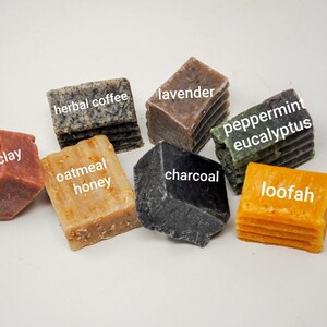 May include: Assortment of eight different bar soaps, each with a unique scent and color. The soaps are arranged on a white surface. The scents are: rose clay, oatmeal honey, herbal coffee, lavender, charcoal, peppermint eucalyptus, and loofah.