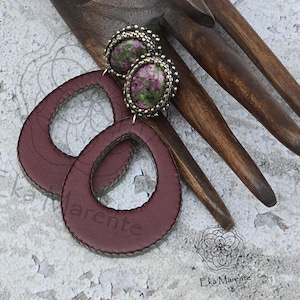 Może przedstawiać: Para kolczyków w kształcie łezki z bordowej skóry i kolczyki sztyfty z zielonymi i różowymi kamieniami oraz srebrnym obszyciem z koralików. Kolczyki są prezentowane na drewnianej dłoni.