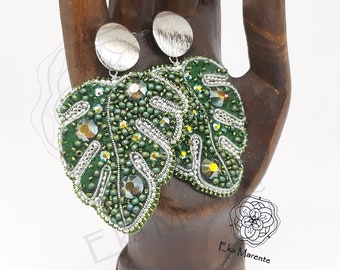 May include: A pair of green monstera leaf earrings with silver beads and rhinestones. The earrings are hanging from silver circular posts.