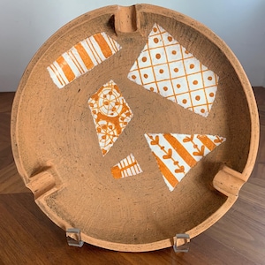 Op de afbeelding: Een ronde asbak van terracotta-kleurige klei met drie geïntegreerde sigarettensteunen langs de rand. Het binnenoppervlak is versierd met verschillende witte en oranje geometrische vlakken, waaronder strepen, ruiten en bloemmotieven.