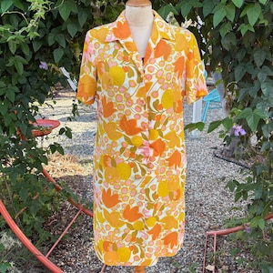 May include: A short-sleeved dress featuring a vibrant floral pattern in shades of orange, yellow, and pink on a white background. The dress has a collared neckline, a button-down front, and a straight silhouette that falls to the knee.