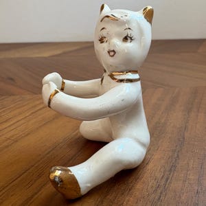 May include: A white ceramic figurine of a seated figure with cat-like ears, featuring gold-painted accents on the ears, neck, wrists, and feet. The figure has painted facial features including eyes, eyebrows, and a small mouth, with arms extended forward.