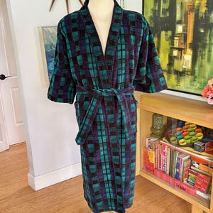May include: A long-sleeved robe featuring a dark blue, black, and teal plaid pattern. The garment includes a matching fabric belt tied at the waist, a shawl collar, and a knee-length hemline, displayed on a mannequin torso for clear visibility.