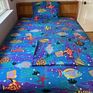 May include: A bed sheet set featuring a vibrant blue background with a colorful pattern of various tropical fish, starfish, and sea plants. The design includes yellow, orange, red, and purple aquatic life scattered across the fabric surface.