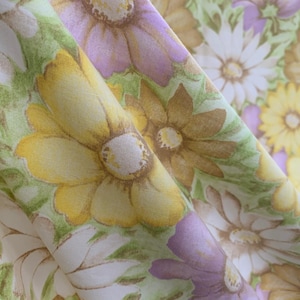 Puede incluir: Primer plano de tela con estampado floral. El diseño presenta grandes margaritas en tonos amarillos, blancos y morados, sobre un fondo verde. La tela está doblada, mostrando la repetición y la textura del patrón.