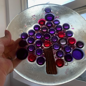May include: A decorative glass plate with a tree design. The tree is made of purple and red glass beads, with a brown trunk. The plate has a silver-coloured, textured background.
