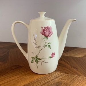 May include: A white ceramic coffee pot with a gold trim and a floral design featuring pink roses and green leaves.