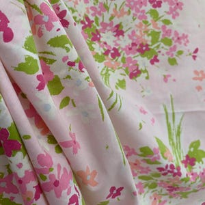 May include: Pink fabric with a floral pattern. The design features clusters of pink, fuchsia, and white flowers with green leaves. The fabric is draped, showing the texture and folds of the material.