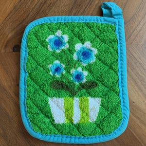 May include: A green quilted pot holder with a blue trim and hanging loop. The pot holder features a floral design with blue and white flowers in a white and green striped pot. The pot holder is approximately 7 inches square.